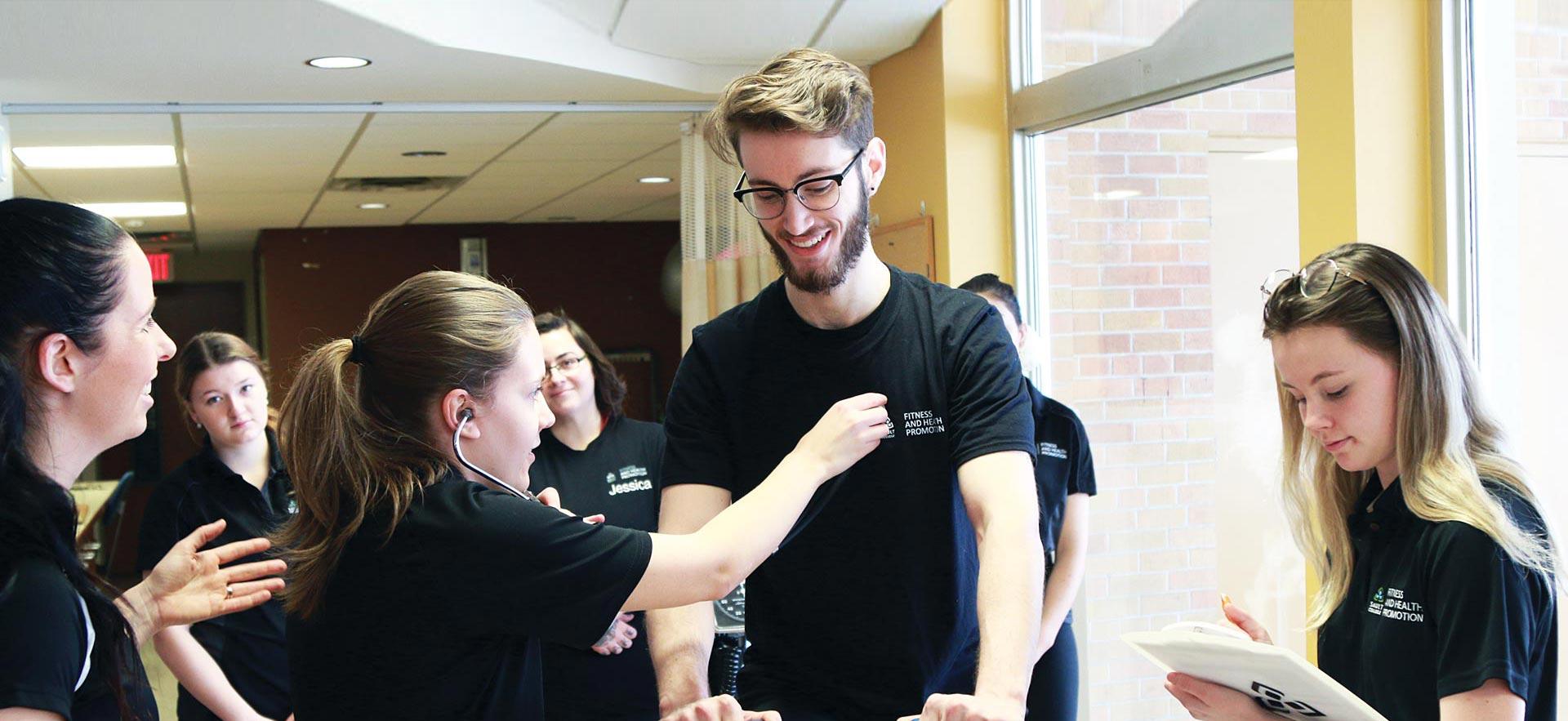 A female Fitness and Health Promotion student checks the  heart-rate of a smiling male student.